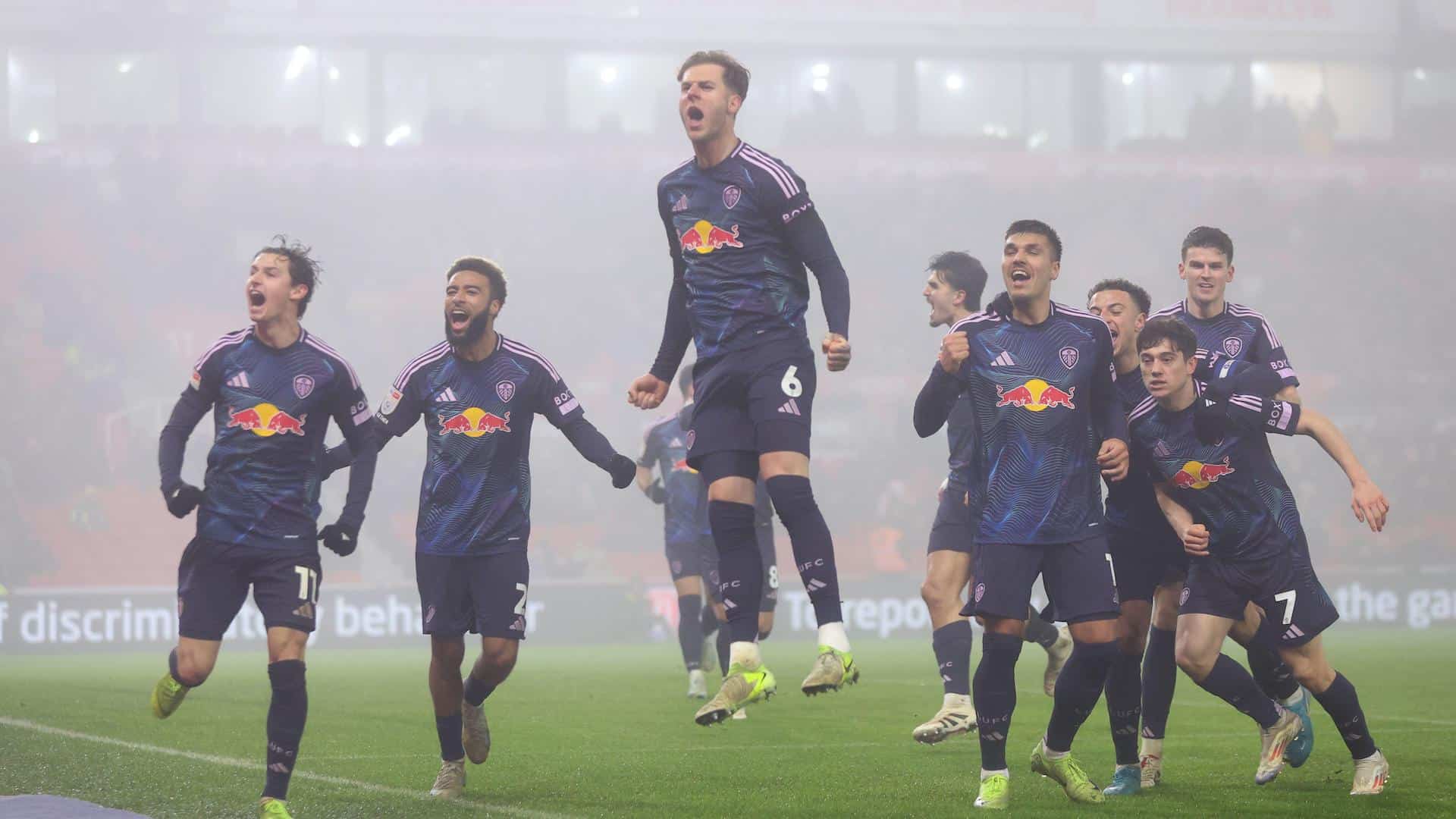 The Leeds United team running out of the fog and towards the away end at Stoke, celebrating Joel Piroe's second goal. Joe Rodon is leading the way, leaping into the air