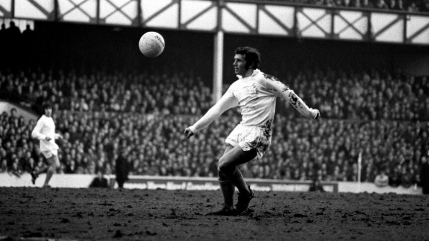 A black and white photo of Johnny Giles dictating play on a pitch that is more mud than grass, with mud covering his white Leeds shirt
