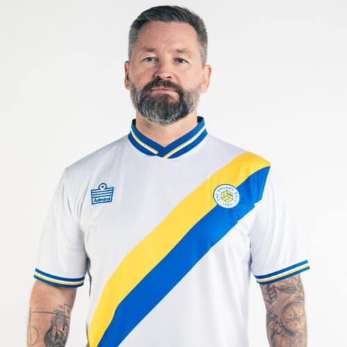 A photo of hunky Svein wearing our 2024 TSB x Admiral home shirt, which is all-white with a blue and yellow diagonal sash