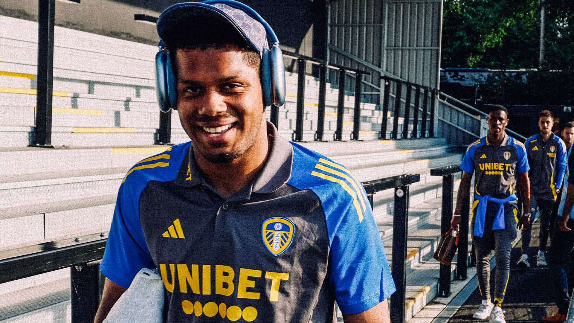 A photograph of Georgi Rutter arriving for Leeds' pre-season friendly wearing headphones over his baseball cap. He looks absolutely delighted, obviously
